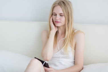 Yatakta otururken kulaklık takıp huzur içinde müzik dinleyen mutlu genç bir kadın..