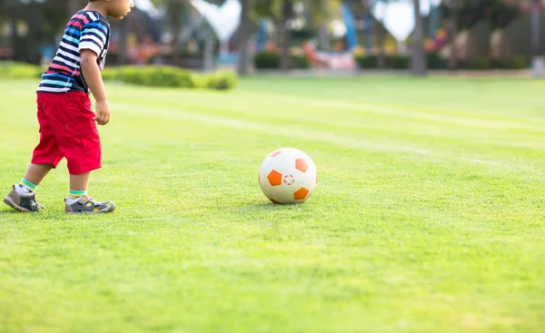 Küçük çocuk futbol oynamayı öğreniyor. Bir yaz günü çimlerin üzerinde topa vurmaya hazırlanıyor..