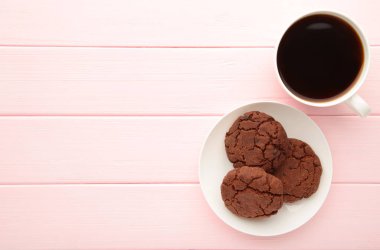Pembe ahşap masada bir fincan kahve ve çikolatalı kurabiye. Üst görünüm