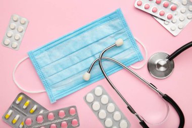 Tıbbi steteskoplu kompozisyon, tek kullanımlık yüz maskeleri, pembe arka planda haplar. Üst görünüm.