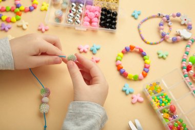Küçük kız bej arka planda bilezikler yapmış. El yapımı boncuklu mücevherler. Kolyeler ve bilezikler çok renkli boncuklar ve incilerden yapılmış. Bilezik boncukları..