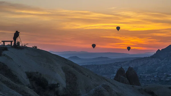 Photographer with tripod on hill makes a shot of hot-air balloon flying over sunrise Cappadocia.