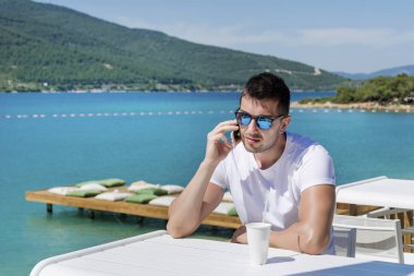 Güneş gözlüğü takan adam, deniz kenarındaki kafede akıllı telefonda konuşuyor
