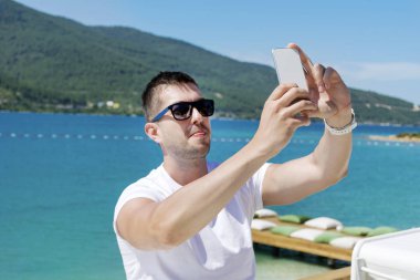 Güneş gözlükçü adam deniz kenarında akıllı telefonda selfie çekiyor