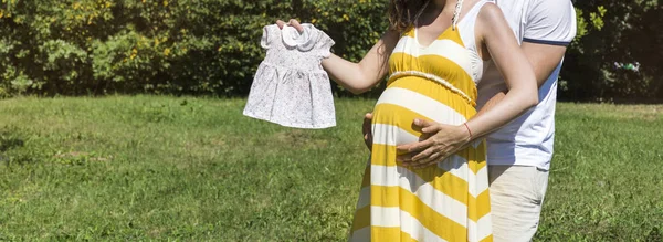 happy family expecting baby  holding baby's clothes  in a summer park