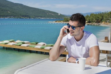 Güneş gözlüğü takan adam, deniz kenarındaki kafede akıllı telefonda konuşuyor