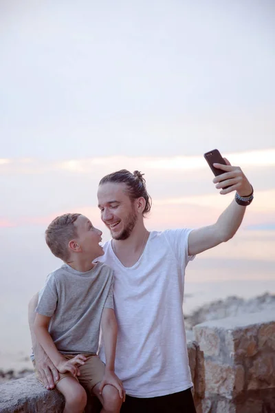 Cheerful bearded dad and little son talking selfie at summer sea sunset. Summer and traveling concept. Copy space.