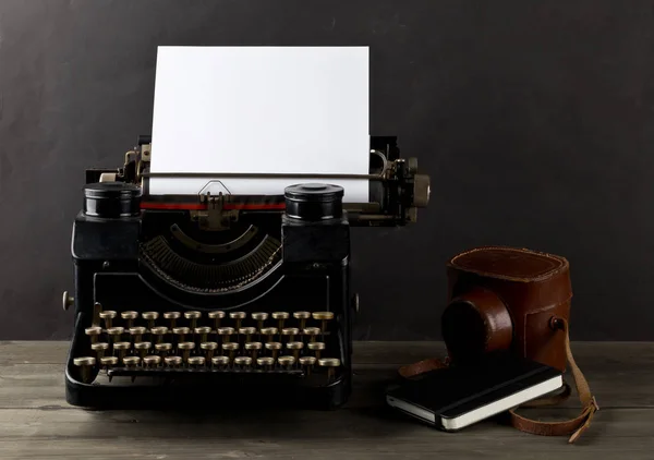 Vintage typewriter with empty, blank sheet of paper, camera and notebook on wood table with black background