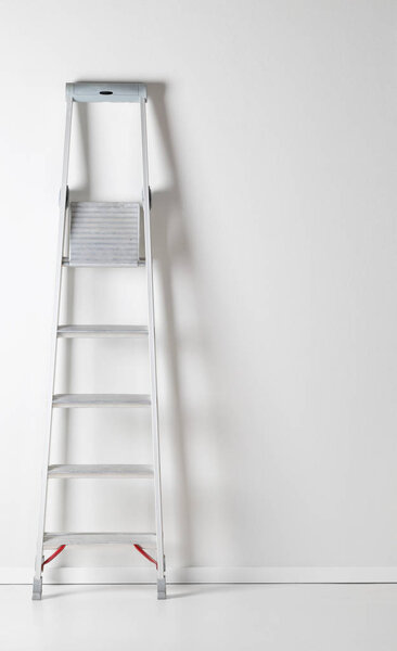 Single aluminum folding metal step ladder leaning against white plaster wall background