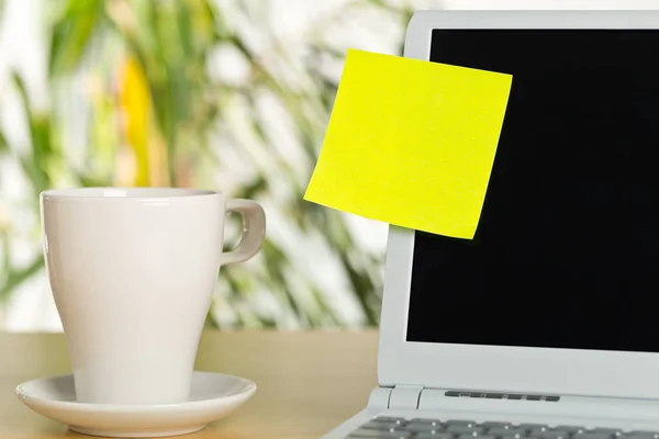 Yellow empty sticky note on laptop screen with white coffee or tea cup with copy space on brown wooden desk in office - selective focus