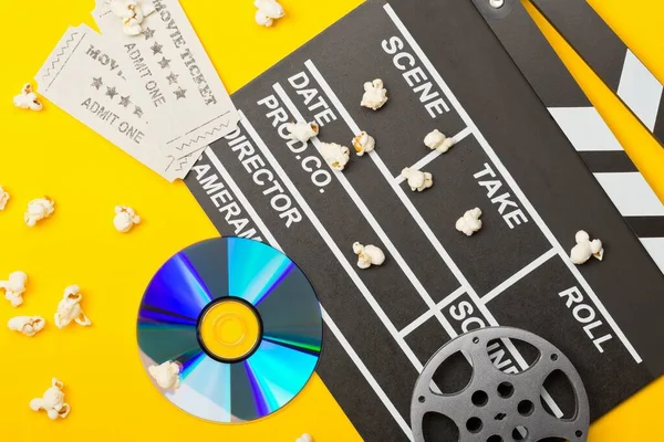 Single, black, open movie clapper or clapper-board with dvd movie disc, film reel, popcorn and movie theatre tickets flat lay top view from above on yellow or orange background - digital movie, home cinema or movie night concept