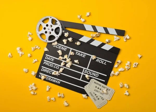 Single, black, open movie clapper or clapper-board with film reel, popcorn and movie theatre tickets flat lay top view from above on yellow or orange background - home cinema or movie night concept