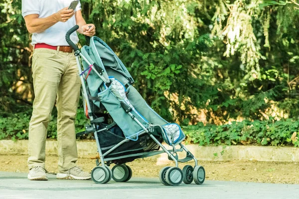 Padre joven con smartphone esperando con carrito de bebé en el parque —  Fotos de Stock