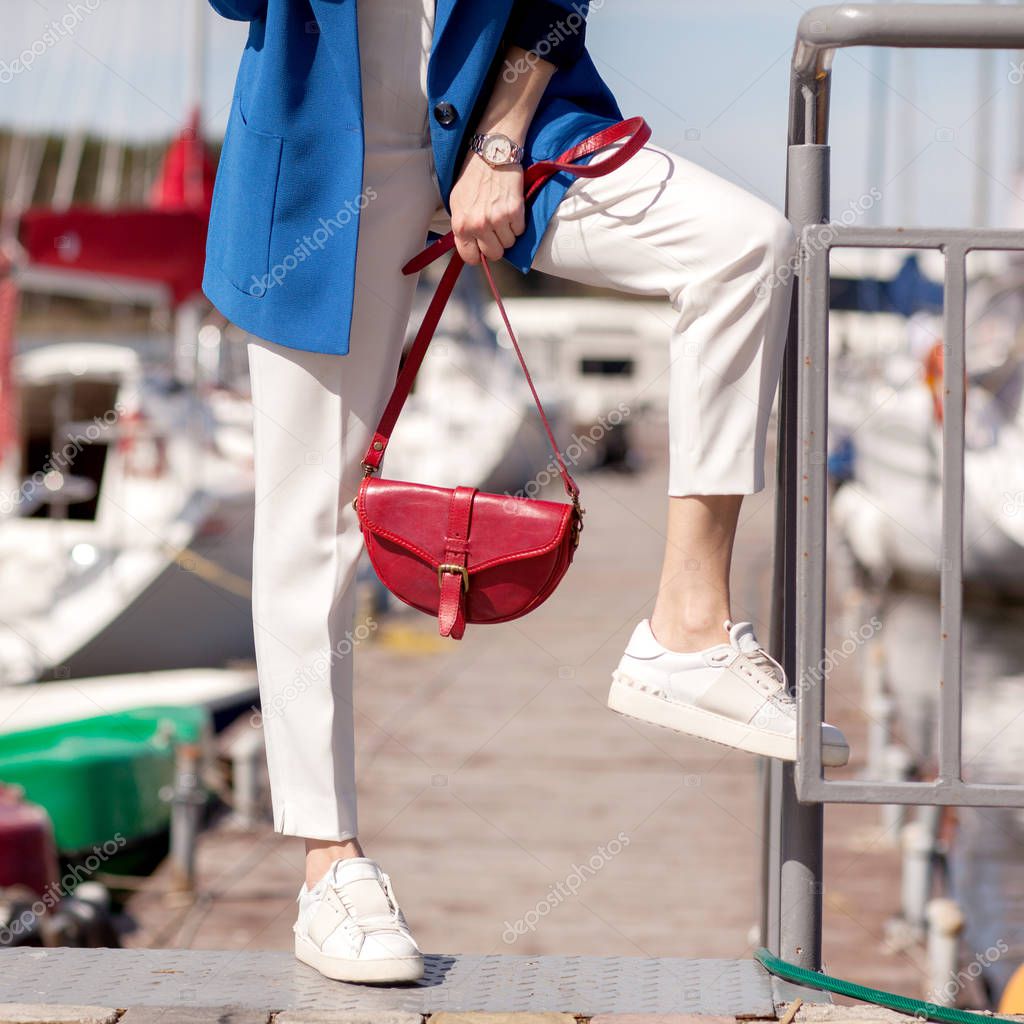 Street, bright style. Young woman in a blue jacket, white sneakers, white trousers, a red handbag, Sunglassesand and a red case for sunglasses. Details. Sguare image photo