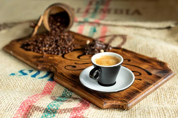 Grãos de café espalhados, uma xícara de café expresso, pedaços de chocolate com nozes em uma tábua de madeira . — Fotografia de Stock