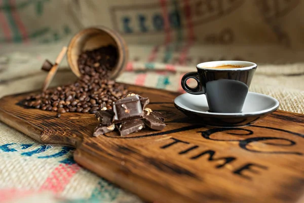 Grãos de café espalhados, uma xícara de café expresso, pedaços de chocolate com nozes em uma tábua de madeira . — Fotografia de Stock