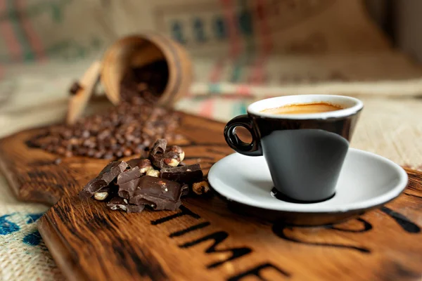 Grãos de café espalhados, uma xícara de café expresso, pedaços de chocolate com nozes em uma tábua de madeira . — Fotografia de Stock