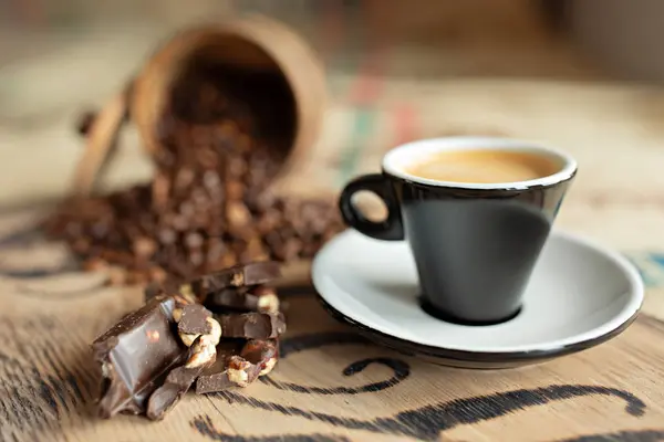 Grãos de café espalhados, uma xícara de café expresso, pedaços de chocolate com nozes em uma tábua de madeira . — Fotografia de Stock