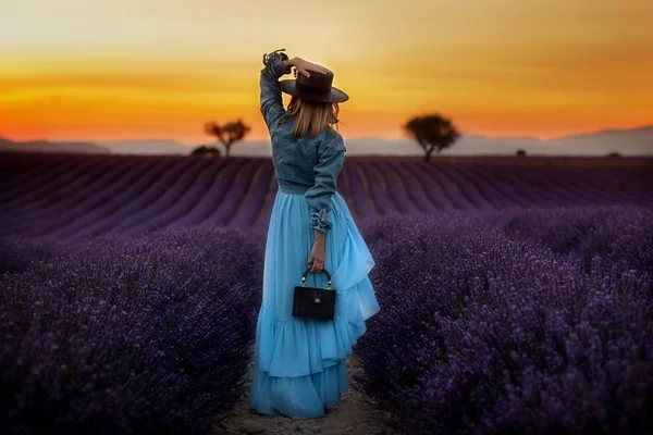 Menina elegante em um vestido suavemente azul e um chapéu preto fica em um campo de lavanda e olha para um belo pôr do sol . — Fotografia de Stock