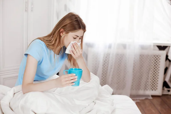 A sick girl is sitting under a blanket on a bed in a bedroom, holding a paper handkerchief, blowing her nose, experiencing relief of nasal congestion and drinking hot medicine. Seasonal diseases, flu, allergies. Space for text