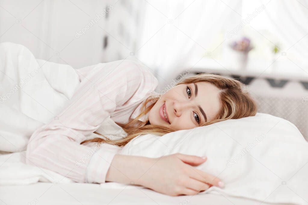 A young attractive girl in striped pajamas is lying in bed in her bedroom in the morning . Side view, space for text