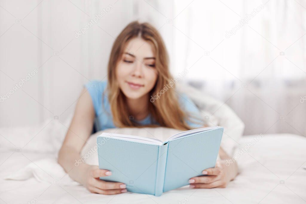 An attractive young girl is lying on the bed in the bedroom and reading a book. Blurred background, space for text