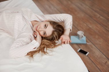 Çekici bir kız yatağa uzanıyor ve neşeyle gülümsüyor, yere yakın bir not defteri, telefon ve bir fincan kahve var. Yukarıdan görünüm, metin için yer