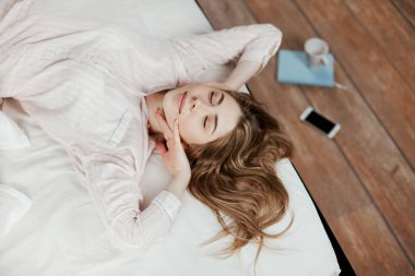 Kapalı gözlü bir kız uyandıktan sonra yatağa uzanıyor ve gülümsüyor, yanında bir defter, telefon ve bir fincan kahve var. Yukarıdan görünüm, metin için yer
