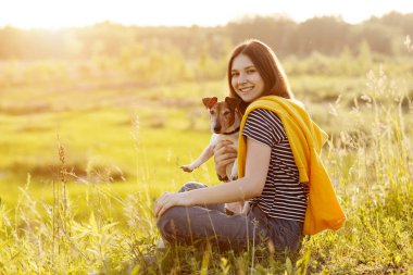 Uzun siyah saçlı güzel bir kız beyaz bir Jack Russell Terrier 'a sarılıyor. Bir kız ve köpeği güneş altındaki çimlerin üzerinde oturuyorlar.