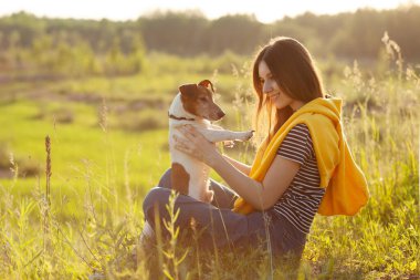 Genç bir kız batan güneşin ışınlarında çimlerin üzerinde oturur ve köpeğini kollarına alır. Köpek gezdirme. Arkadaşlık, dikkat. Yan görünüm, metin için boşluk