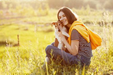 Çekici bir kız çimenlerde oturuyor, gülümsüyor ve köpeğine sarılıyor. Bir kız ve evcil hayvanı temiz havada dinleniyorlar. Arkadaş, dostluk, ilgi