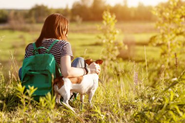 Sırtında yeşil bir sırt çantası ve çizgili tişörtü olan koyu renk saçlı bir kız köpeğe sarılıyor ve çayırdaki çimlerin üzerinde oturuyor. Yürüyüşten sonra bir kız evcil hayvanıyla dinleniyor. Arka plan, metin için boşluk