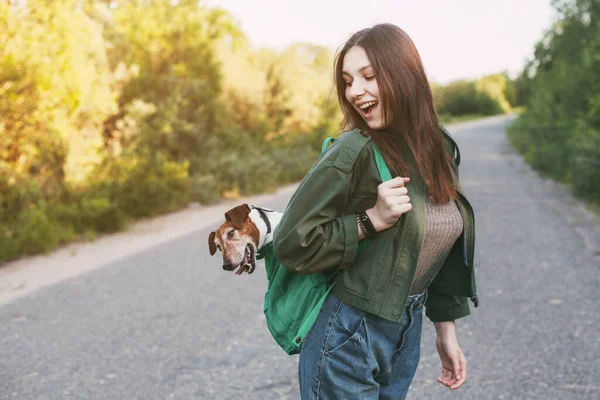 美しい少女が肩に緑のバックパックを持っていて そこから可愛い犬が外を見ている 女の子と彼女の友人が一緒に旅行し 散歩をします 友情とケアの概念 — ストック写真