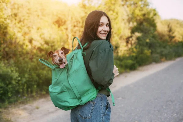 笑顔の女の子が肩に緑のリュックを抱えていて そこから可愛い犬が外を見ている 女の子と彼女の友人が一緒に旅行し 散歩をします 友情とケアの概念 — ストック写真