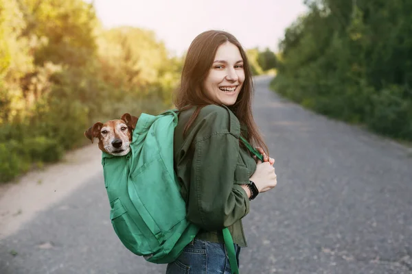 笑顔の女の子が肩に緑のリュックを抱えていて そこから可愛い犬が外を見ている 女の子と彼女の友人が一緒に旅行し 散歩をします 友情とケアの概念 — ストック写真