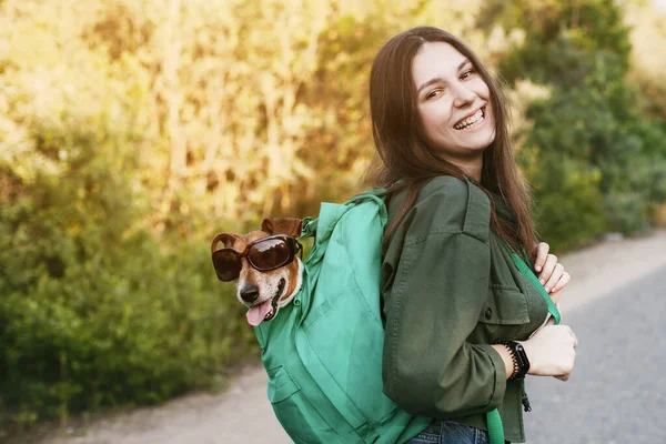 笑顔の女の子が肩に緑色のバックパックを持っていて そこからサングラスをかけている犬が外を見ている 女の子と彼女の友人が一緒に旅行し 散歩をします 友情とケアの概念 — ストック写真