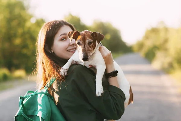 肩に緑のリュックを背負ったジャケットの少女が腕に犬を抱えて抱きかかえている 少女はペットと一緒に歩く リアビュー テキストの場所 — ストック写真