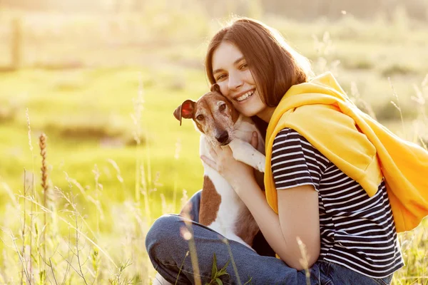 魅力的な女の子は 草の上に座って 彼女の犬を笑って抱きかかえています 新鮮な空気の中で少女と彼女のペットがリラックスしている — ストック写真