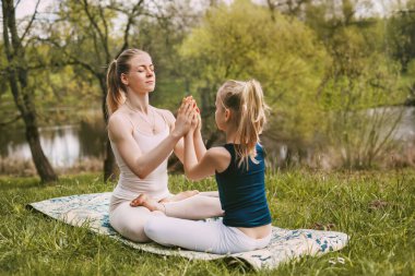 Genç bir kadın ve kızı temiz havada yoga yapıyorlar. Anne ve kızı ellerini birleştirip Lotus pozisyonunda oturuyorlar. Aile tatili, meditasyon. Yan görünüm, metin için boşluk
