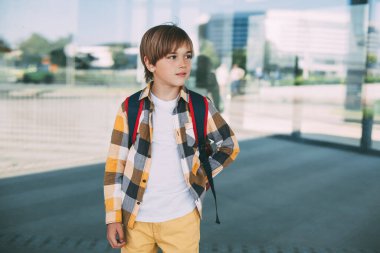 Sırt çantalı mutlu bir çocuk okulun yanında duruyor, güzel gülümsüyor ve düşünceli bir şekilde uzaktan bakıyor. Yaz tatilinden sonra yeni okul yılı başlıyor.