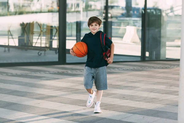Sırt çantası ve basketbol topu olan neşeli bir çocuk. Bir çocuk basketbol antrenmanına koşar. Eğitim, eğitim