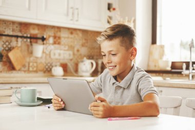 Mutfak masasında tabletle oturup arkadaşlarıyla sohbet eden mutlu çocuk, yakın plan.