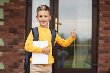 Elinde evrak çantası ve tabletle gülümseyen bir çocuk evinin yanında duruyor. Sınıfı parmağınla gösterir.