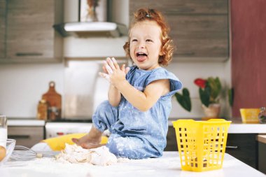 Una bulanmış neşeli küçük bir kız, masaya oturur ve güler, masanın yanında hamur ve mutfak gereçleri, mesaj için yer vardır.