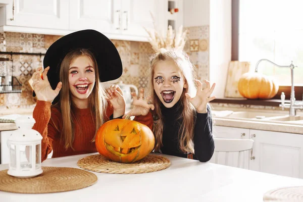 Due ragazze a casa in cucina in costume e trucco per Halloween siedono con una zucca, giocano e ridono. I bambini felici si stanno preparando per Halloween. — Foto Stock