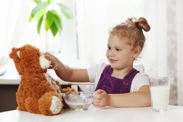 Una ragazza sta giocando al tavolo e sta dando cereali al suo amico orsacchiotto. La ragazza mangia cereali con latte per colazione. Colazione sana, prendersi cura dei bambini. Spazio per testo — Foto Stock