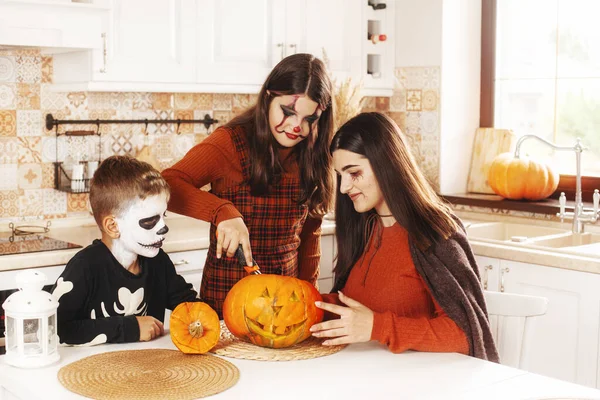 Zusters en broer zijn thuis in de keuken in kostuums en make-up voor Halloween, snijden een pompoen, spelen en lachen. — Stockfoto