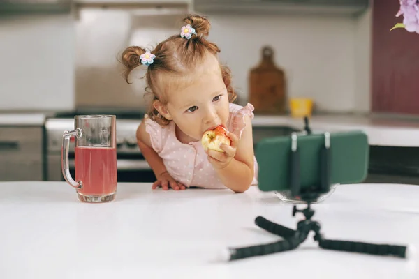 Ritratto di una graziosa bambina seduta al tavolo della cucina, che mangia una mela e guarda un video sul suo telefono — Foto Stock