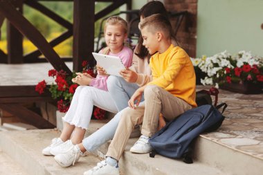 Okuldan sonra bir grup mutlu genç çocuk basamaklarda oturup dijital tablette oynuyorlar. Modern çocuklar, dijital teknolojiler