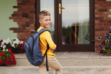 Elinde evrak çantası ve tabletle gülümseyen bir çocuk evinin yanında duruyor. Okula gitmeye hazır. Okula geri dön..